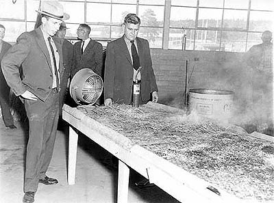 In a Forestry Department warehouse, foresters observe a demonstration of a simulated burn: a raised tray of grass burns and smolders as an electrical fan mounted to one end generates a simulated breeze.