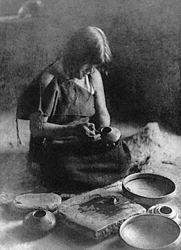 Image from 1906: a young Yavapai woman in traditional dress sits on the floor of her studio, hand detailing a small pot.