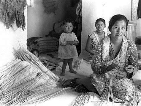 Image from circa 1946: two generations of Hopi women in simple cotton print dresses are sitting on the floor of their home, splitting needles by hand while a toddler in a shirt watches.