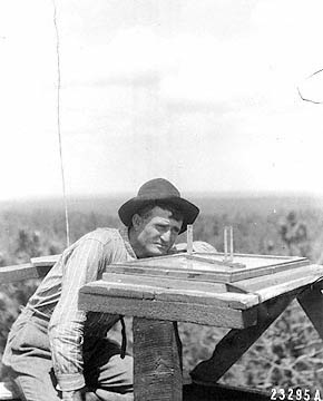 Atop a tower well above the trees, a lookout in his late forties crouches at a surveying table to sight a distant fire.