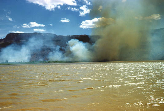 Seen from across a river on a bright sunny spring afternoon in 1949, plumes of white and brown smoke from burning brush obscure the mesas in the distance.