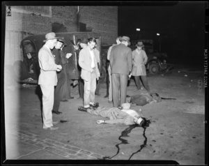 A crime scene, showing a number of people standing around a body, next to a car.