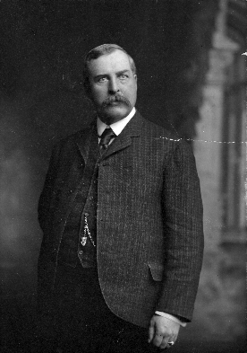 A heavyset, middle-aged man poses for the camera, wearing a suit and vest, with a heavy moustache and a receding hairline. His coat is unbuttoned and he has a determined expression, head partially turned to one side.