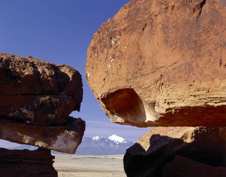 Image depicts the San Francisco Peaks in distance through large stacked sandstone rocks in the foreground. The sky is clear and lighting direction is from above right.