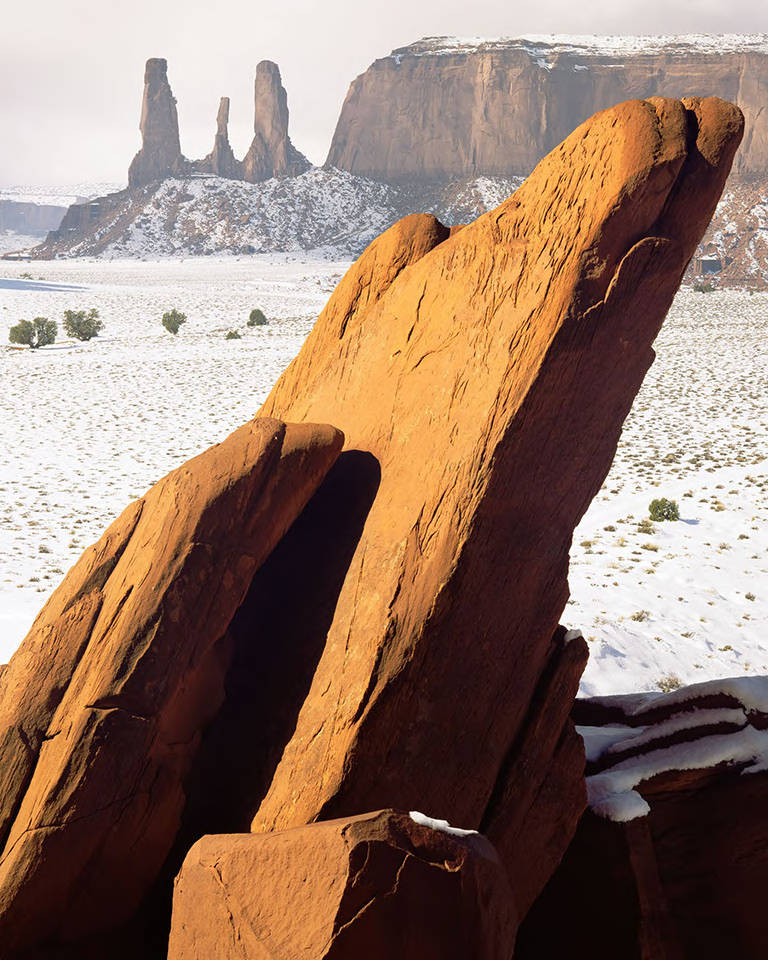 This image shows a sandstone rock in the foreground, with the Three Sisters rock formation in Monument Valley, Arizona in the background. Snow is visible on the ground.